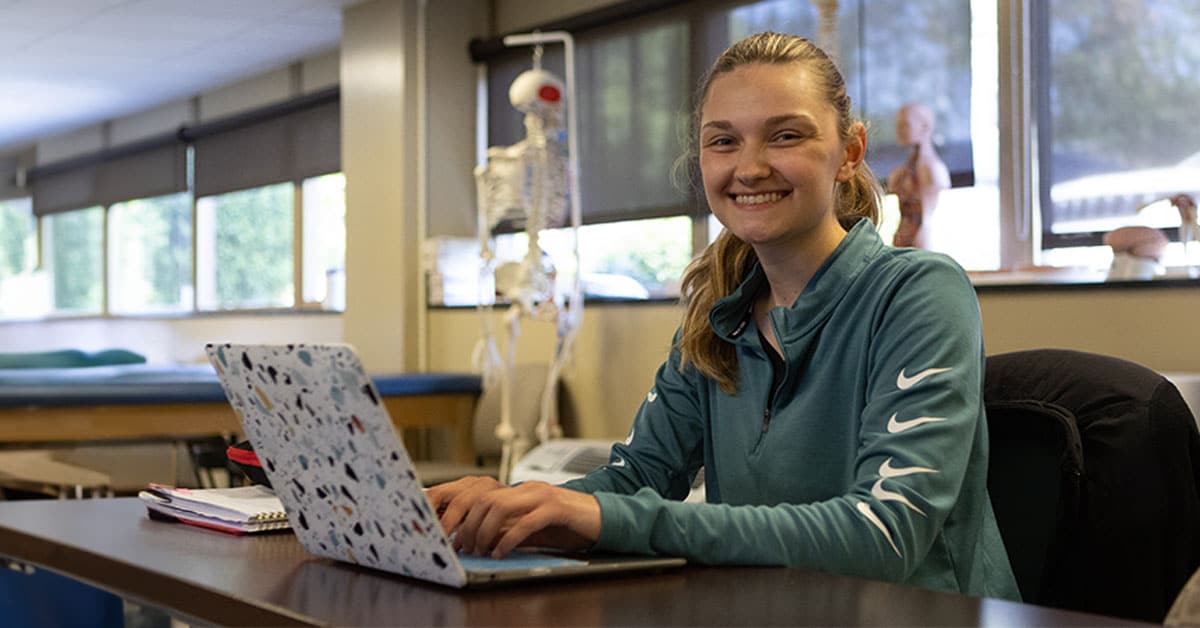 A young woman smiles while typing on a laptop in a classroom with anatomical models in the background.