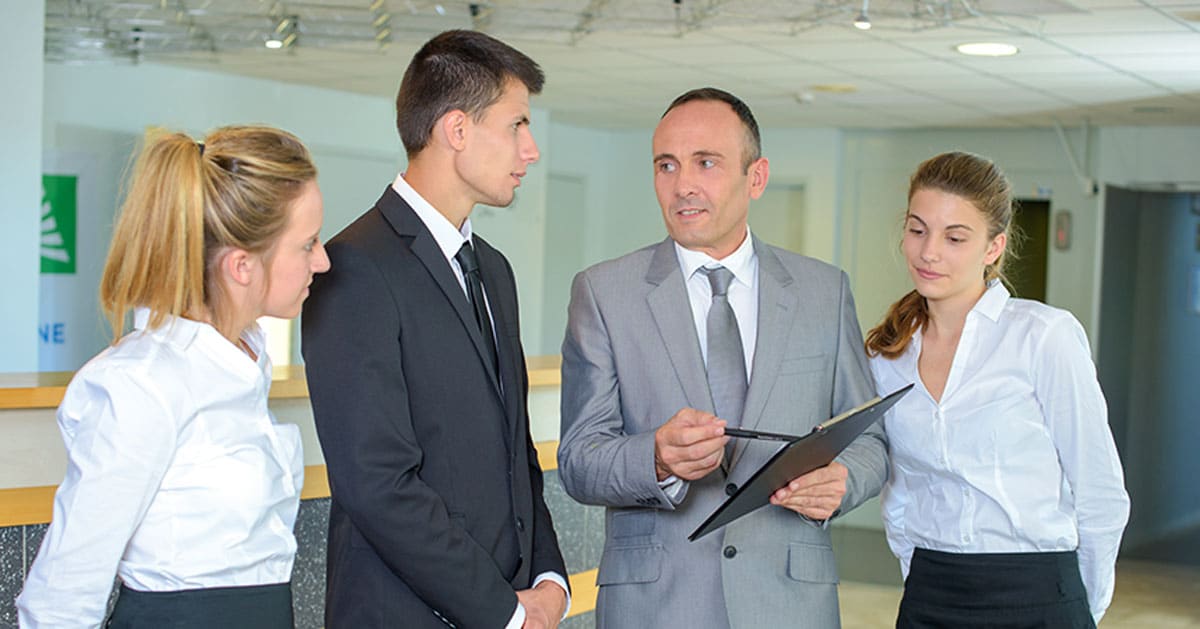 A man in a grey suit discussing something on a clipboard with three colleagues in business attire.