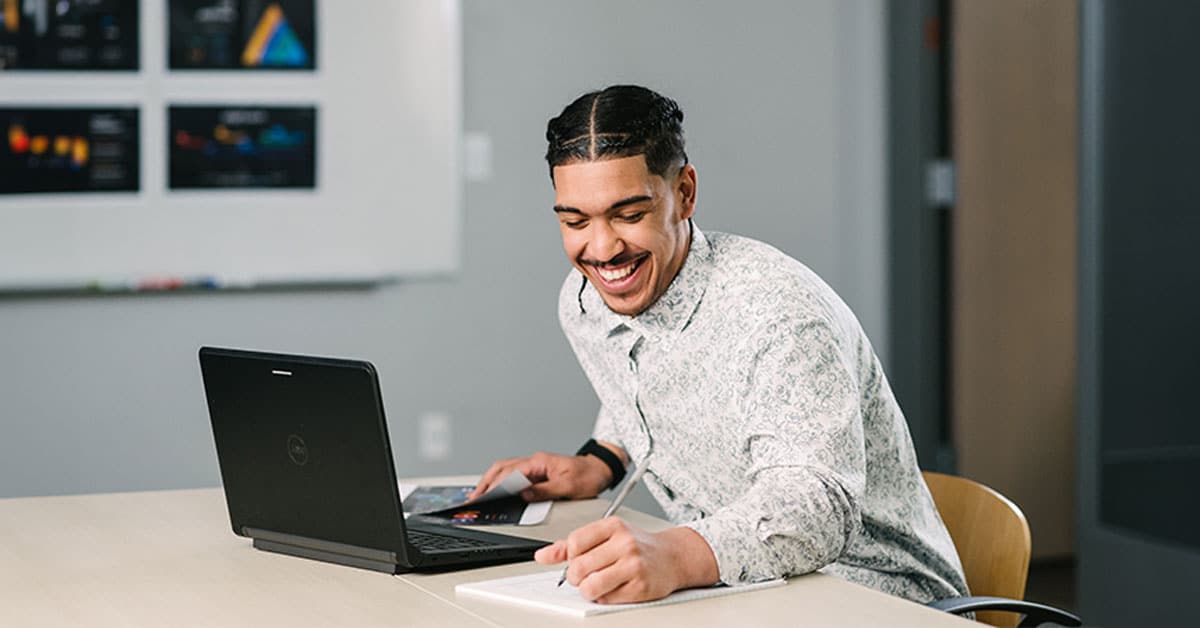 A person smiling while using a laptop and writing in a notebook at a desk in an office setting.