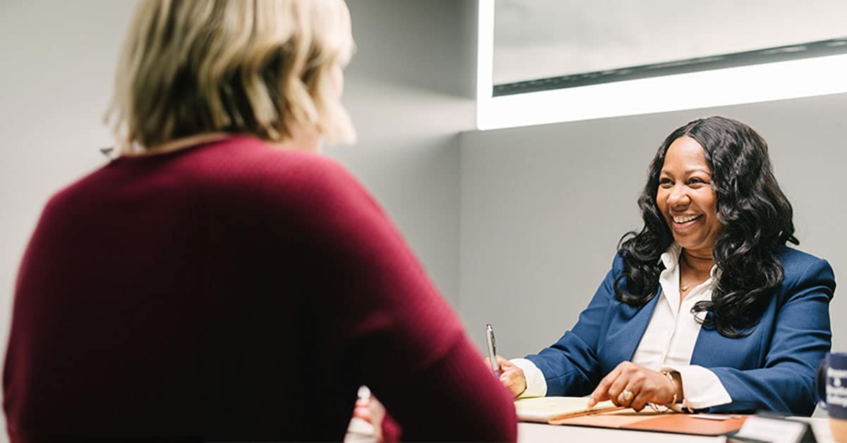 Two women are sitting across a table in an office, engaging in a friendly conversation.