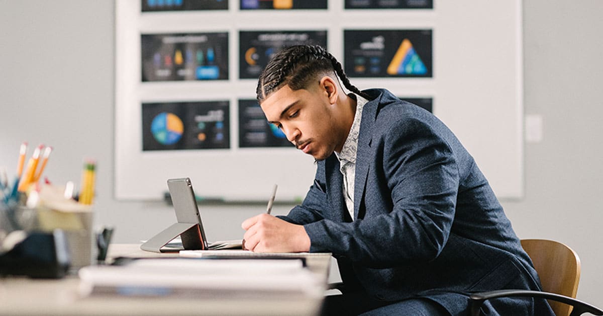 A man in a suit writes notes on paper, using a tablet for reference, with charts on a whiteboard in the background.
