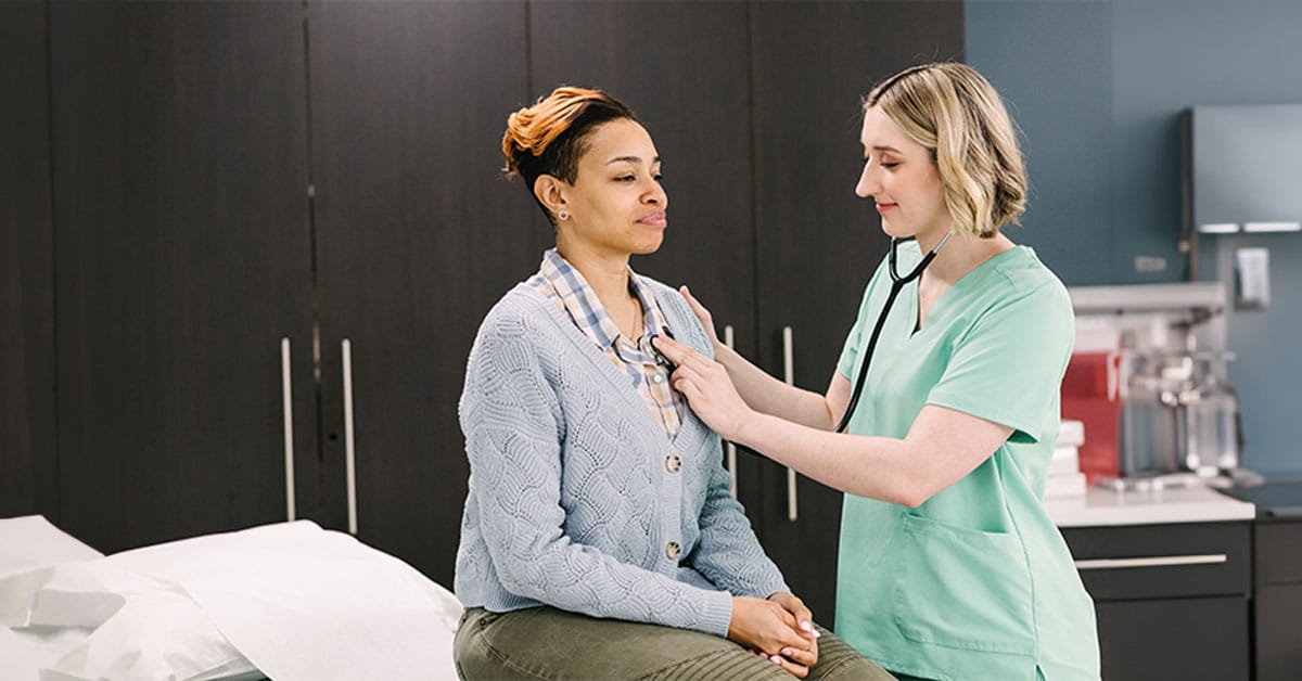 A woman in a blue sweater sits on an exam table while a healthcare professional in scrubs listens to her chest with a stethoscope.
