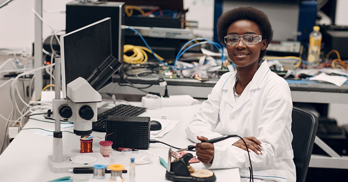 A scientist in a lab coat and safety glasses smiles while working at a cluttered lab desk with electronic equipment.