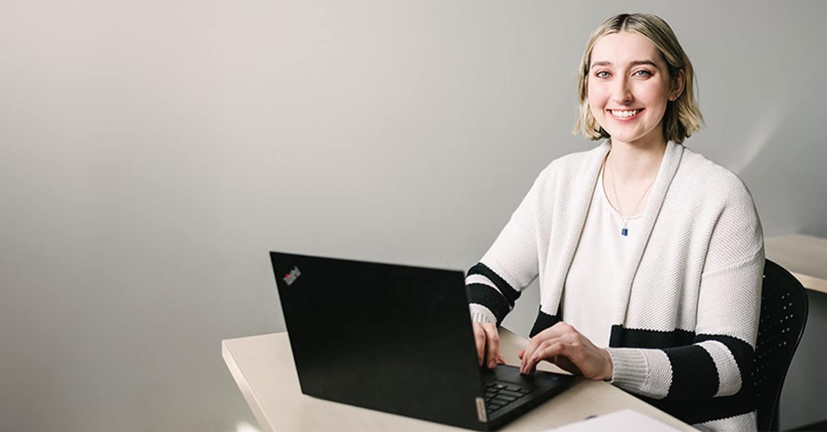 A person with short hair and striped sweater smiles while typing on a laptop at a desk.