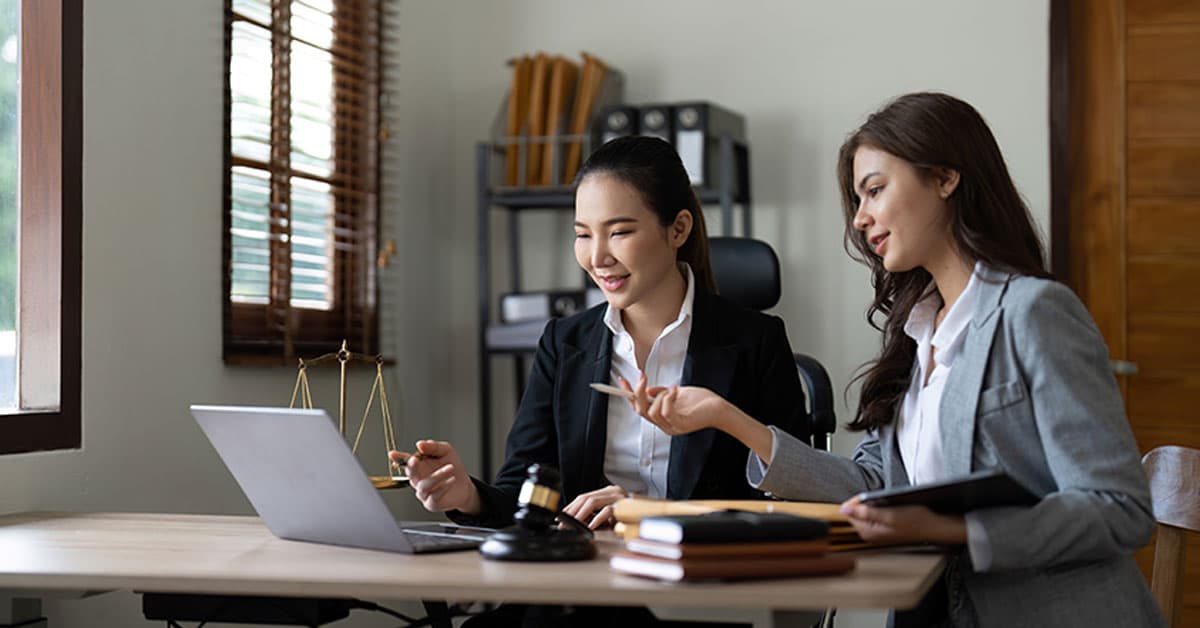 Two women in professional attire discuss work at a desk with a laptop, stack of books, and a gavel in an office.