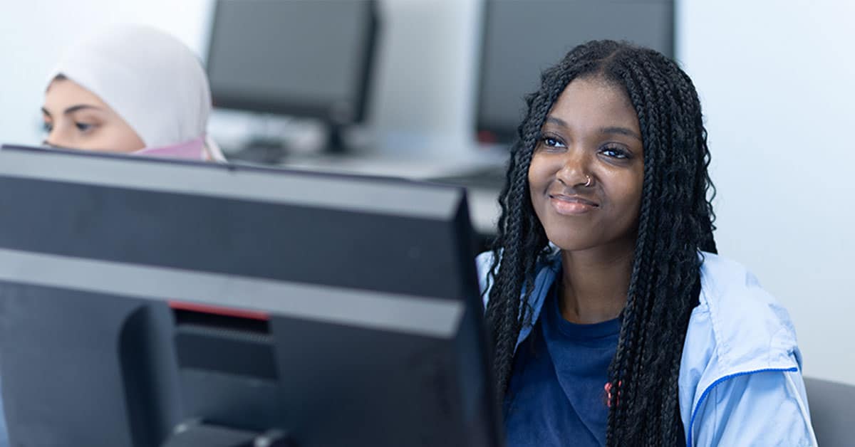 Two women sitting at computer desks, one wearing a hijab and the other with braids, both focused on their screens.