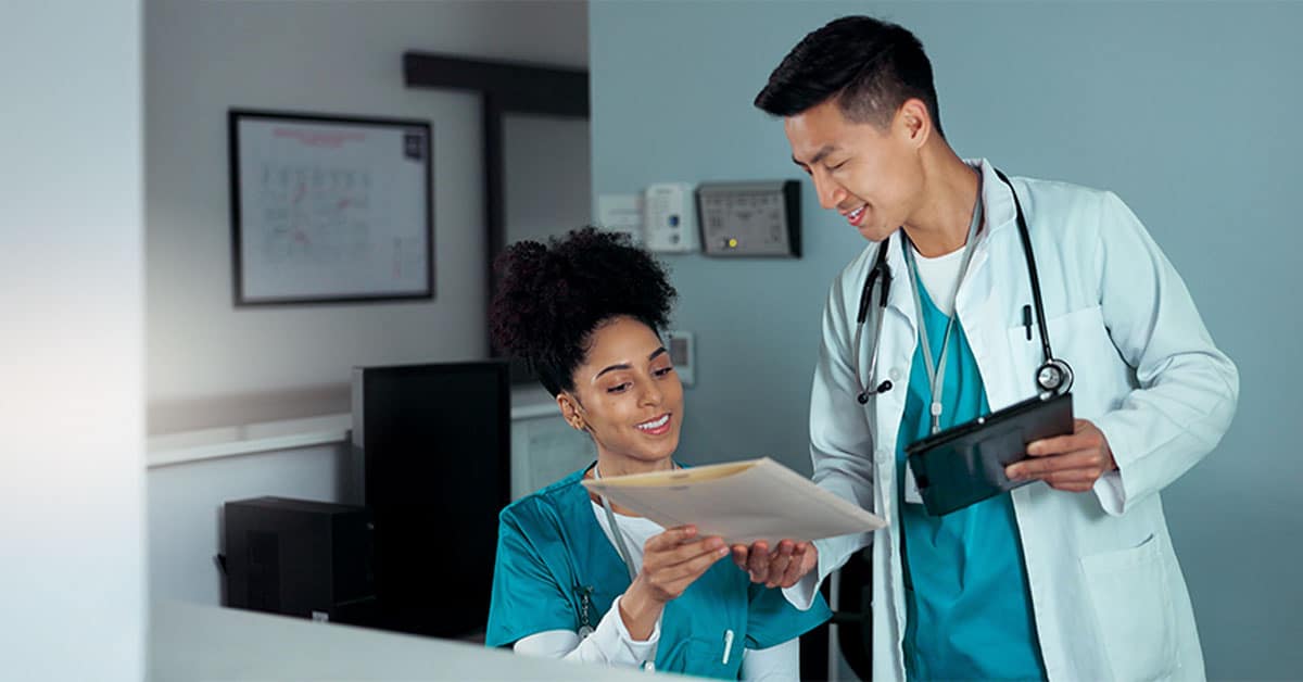 Two healthcare professionals in scrubs discussing patient files; one holds a tablet, the other holds a manila folder.