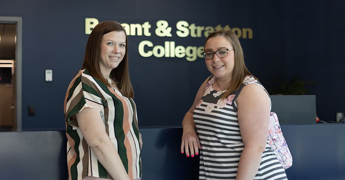 Two women standing and smiling in front of a wall sign that reads "Bryant & Stratton College.
