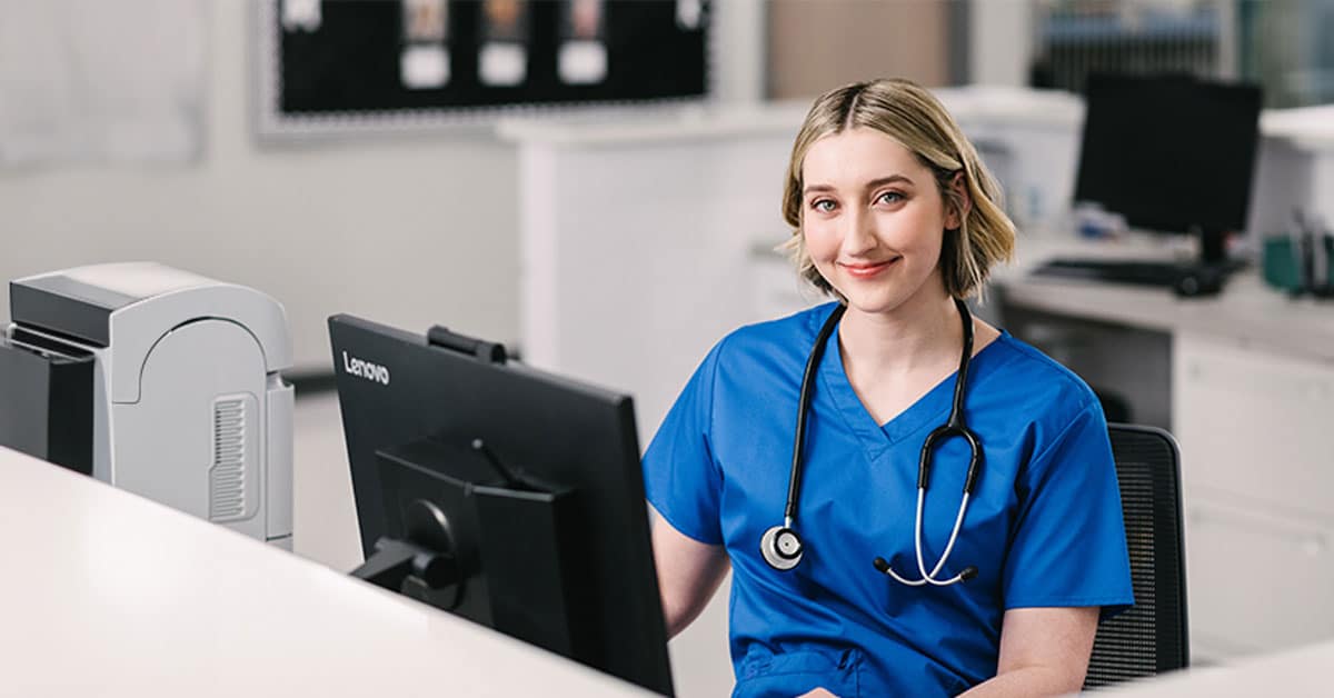A healthcare professional in blue scrubs, sitting at a desk with a computer, smiles warmly.