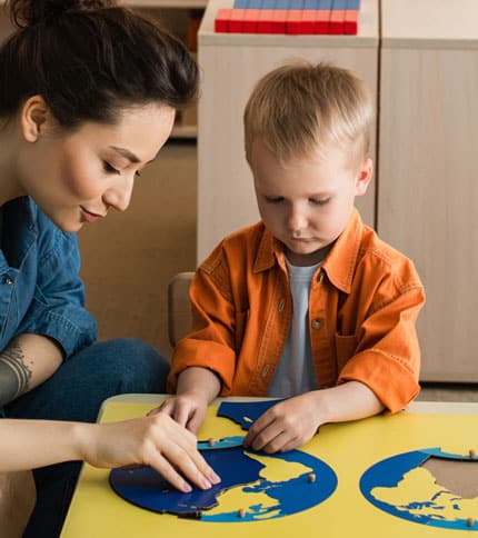 A woman and a young boy are sitting at a table working on a puzzle featuring continents. The boy is focused on placing a piece correctly. Both are engaged in a learning activity, with shelves and books in the background.