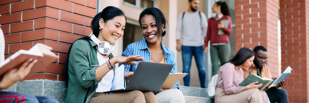 Two students sit on outdoor steps by a brick building, smiling at a laptop. Other students are reading or talking in the background, highlighting the blend of on-campus life with flexible learning opportunities.
