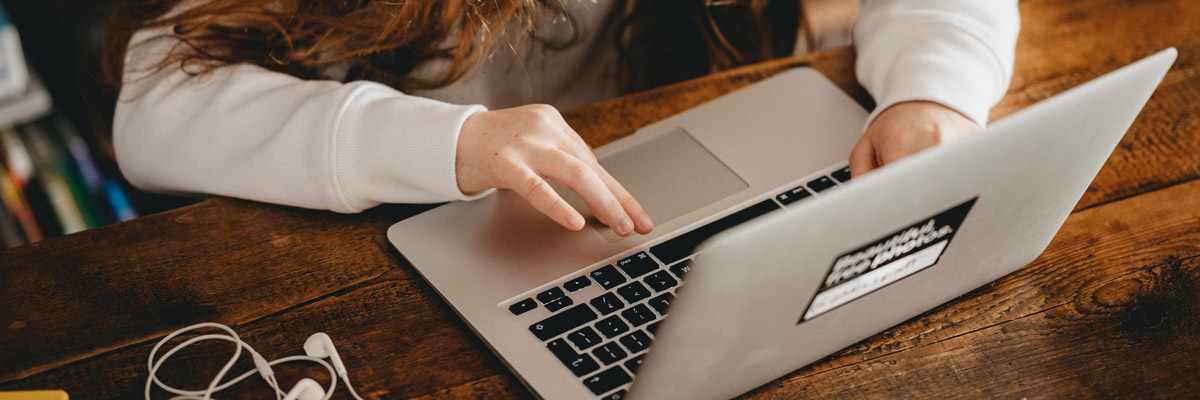 A person with long hair wearing a white sweatshirt uses a laptop on a wooden table, embodying flexible learning. Earphones are lying on the table next to the laptop, suggesting an online learning session in progress.