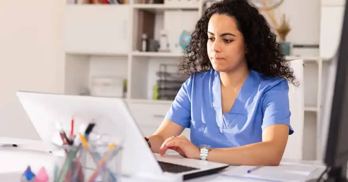 A person with curly hair wearing scrubs uses a laptop at a desk in an office setting.