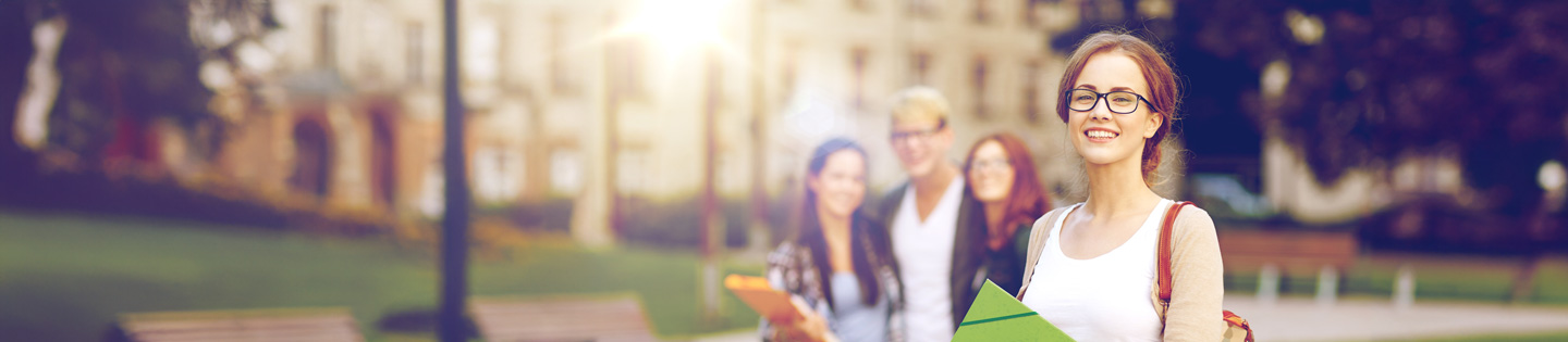A group of students walking on a campus with a woman in glasses smiling and holding books in the foreground.