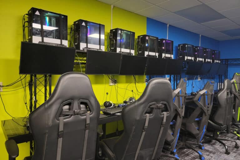 Gaming setup with multiple PCs, monitors, and gaming chairs arranged in a row against a bright yellow and blue wall.