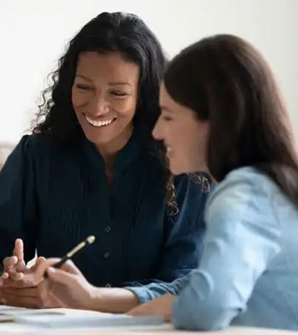 Two women sit together at a table, smiling and talking while looking at documents. One woman is holding a pen, and they appear to be having a pleasant, collaborative conversation.