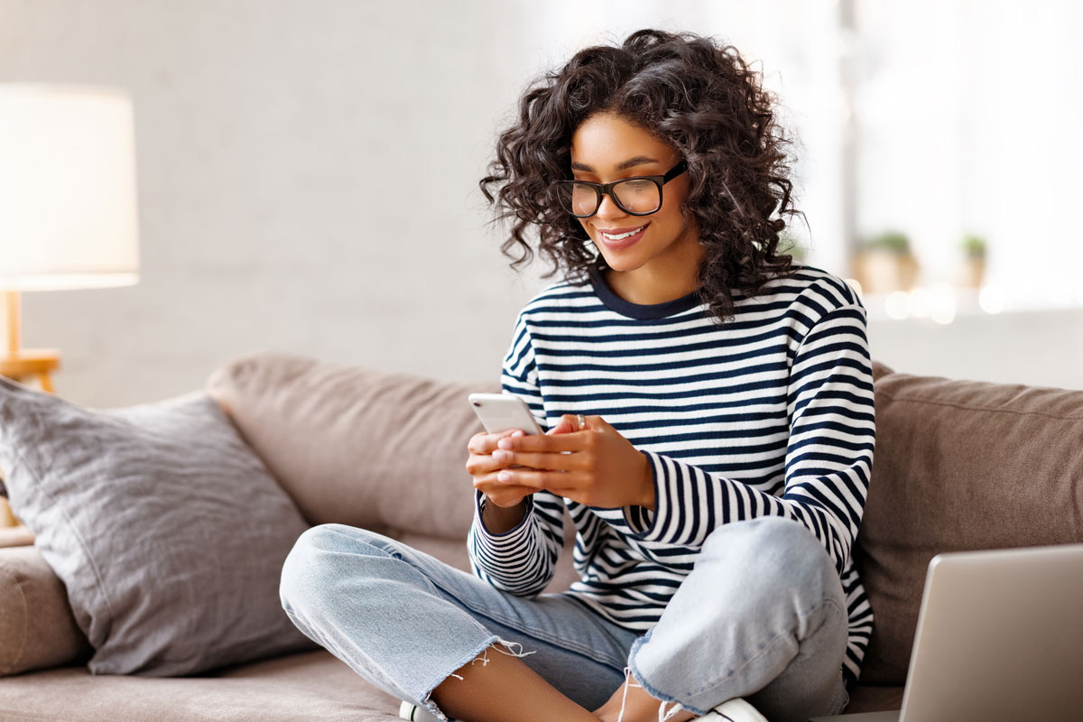 A young woman sits comfortably on a couch, smiling as she uses her smartphone, promoting the ease of downloading the mobile app for seamless access to services on the go.
