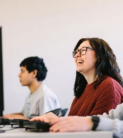 Two people sit at computer desks in a classroom. A woman in glasses and a red sweater is smiling and looking up, while the person next to her focuses on their screen.