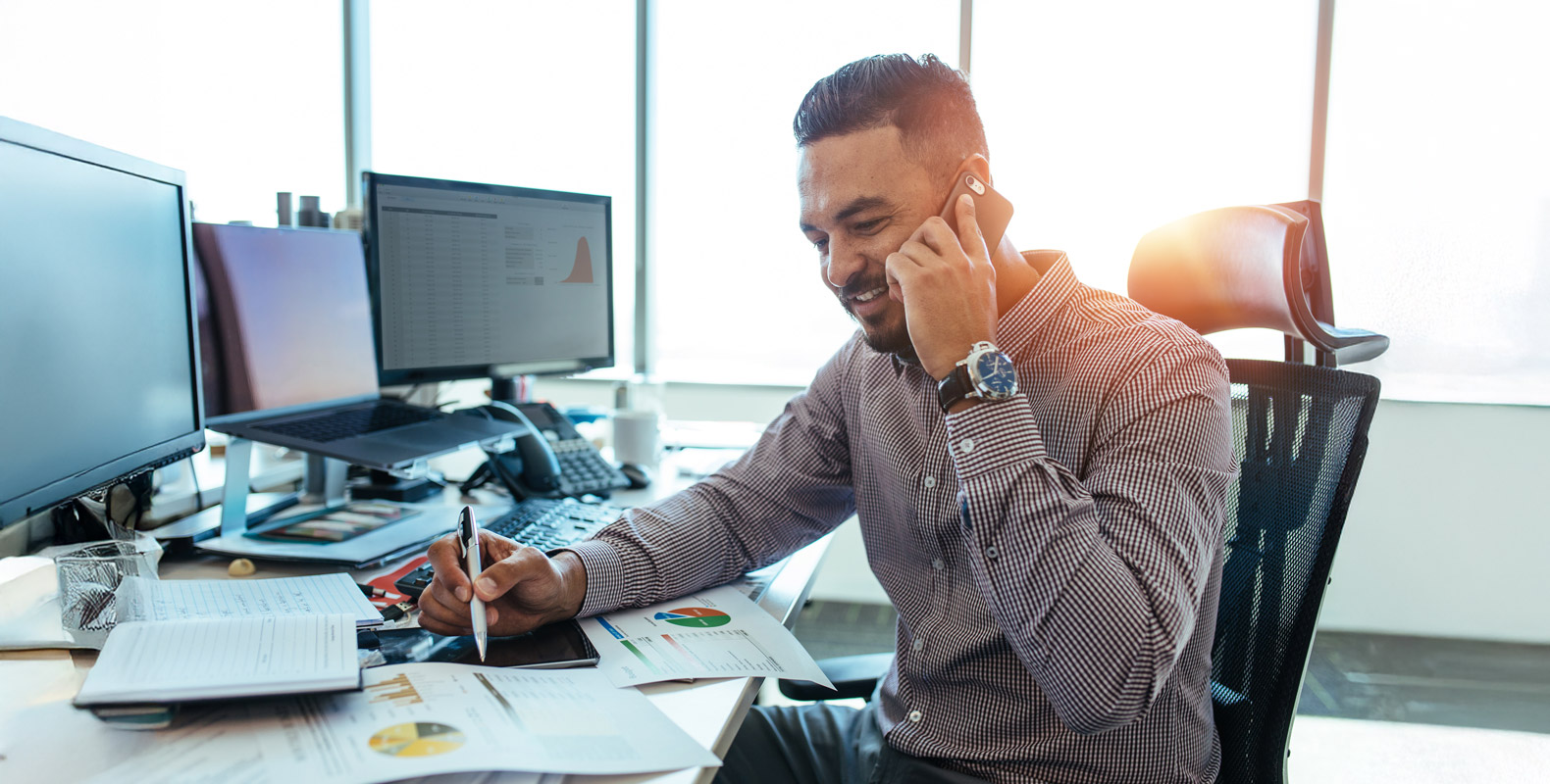 A man in a business casual shirt sits at a desk, talking on the phone while reviewing charts and notes, with multiple computer monitors in the background.