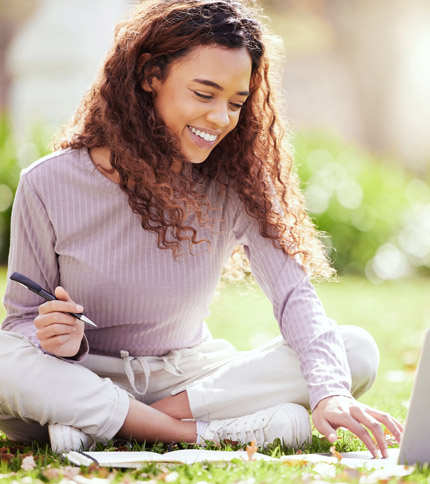 A woman with curly hair sits cross-legged on grass, smiling while writing with a pen on paper. She wears a long-sleeved striped shirt and light pants. Sunlight filters through trees in the background.