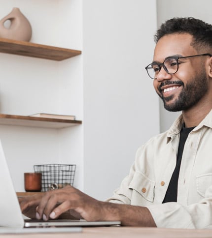 A man with glasses and a beard is smiling while using a laptop. He is seated at a desk in a bright room with wooden shelves and decorative items in the background.