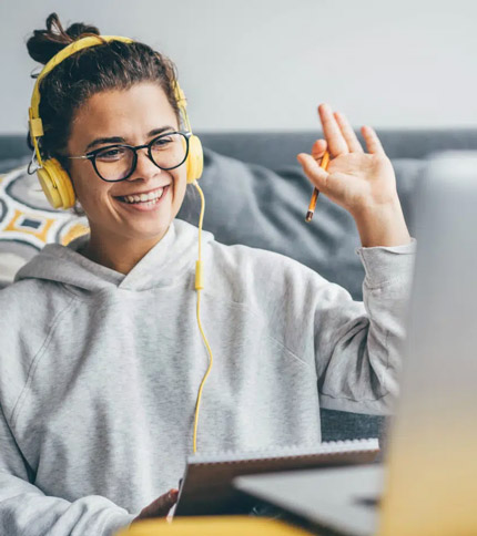 A person wearing glasses and yellow headphones is smiling and waving at a laptop screen. They are seated on a couch, wearing a gray hoodie, and holding a pen. The setting suggests they are participating in a video call or online class.