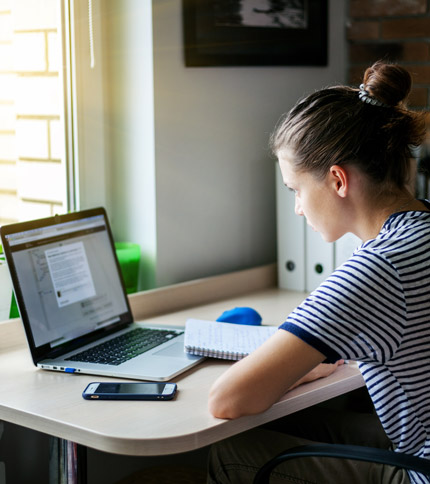 A person in a striped shirt is sitting at a desk, focused on a laptop. They are taking notes in a notebook and a smartphone is placed on the desk. Sunlight is streaming in through a nearby window.