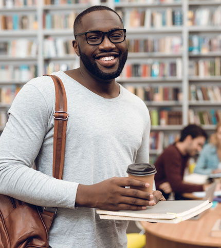 Colleges student in library holding coffee and books with more students and books in the background