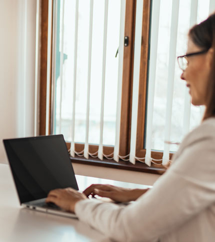A woman sits at a desk, typing on a laptop. She is wearing a white blouse and glasses. Behind her is a window with vertical blinds partially open. The scene suggests she is working in a bright room.