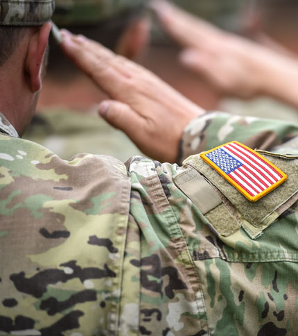 Soldiers in camouflage uniforms saluting, with focus on one soldier's arm displaying a patch of the United States flag.