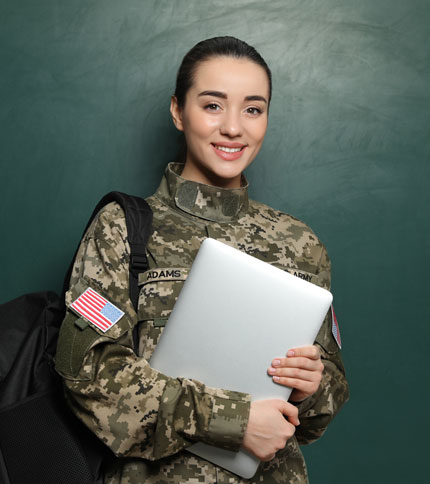 A woman in military uniform smiles while holding a laptop and a backpack. She stands in front of a dark green background. The uniform has an American flag patch and a name tag.