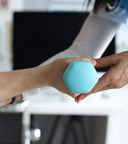 A close-up of a person handing a blue hexagonal dumbbell to another person. The background is blurred, showing a room with exercise equipment. The focus is on the exchange of the dumbbell between the two hands.