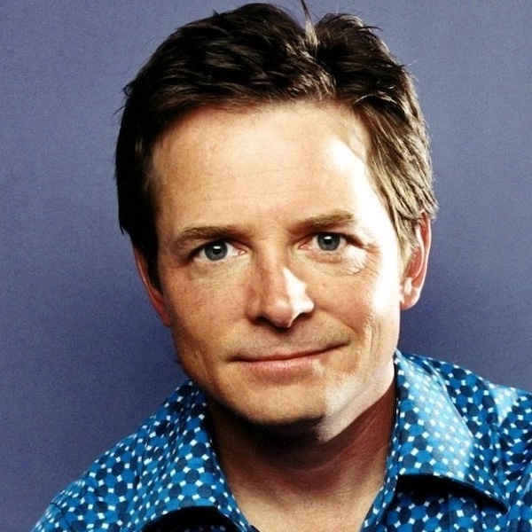 Thomas J. Whitmore, a man with short brown hair and blue eyes, wears a blue patterned shirt and smiles softly at the camera against a plain purple background.