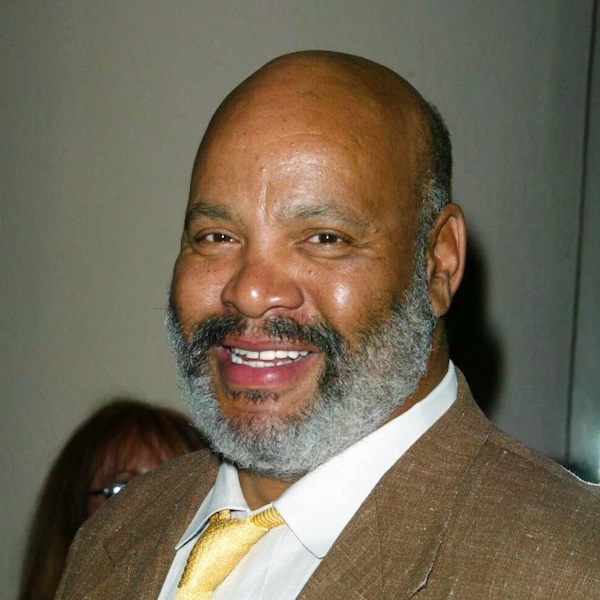 A man with a bald head and gray beard, possibly Thomas J. Whitmore, wearing a brown suit jacket, white shirt, and yellow tie, smiles at the camera. The background is plain with a partially visible person behind him.