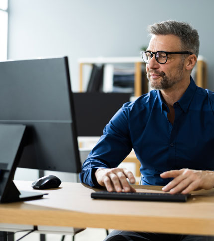 A man wearing glasses and a dark blue shirt is sitting at a desk, typing on a keyboard and looking at a computer monitor. The office background has shelves with books and decor items.