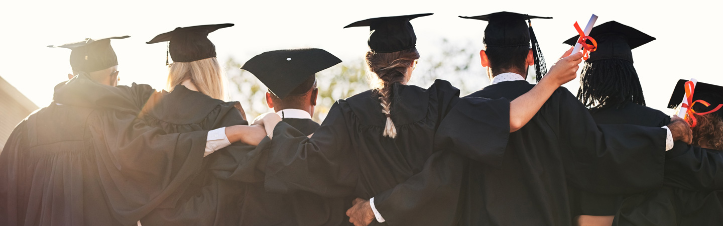 A group of graduates wearing black caps and gowns stand with their backs to the camera, arms around each other. One person holds a diploma tied with a red ribbon. The sky is clear in the background.