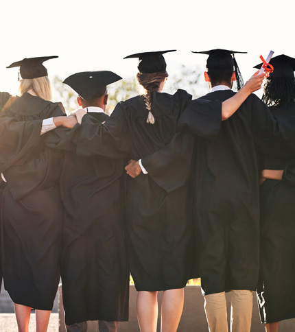 A group of graduates in black caps and gowns stand with their backs to the camera, arms linked, against a bright sky. One holds a diploma, and the scene conveys celebration and achievement.