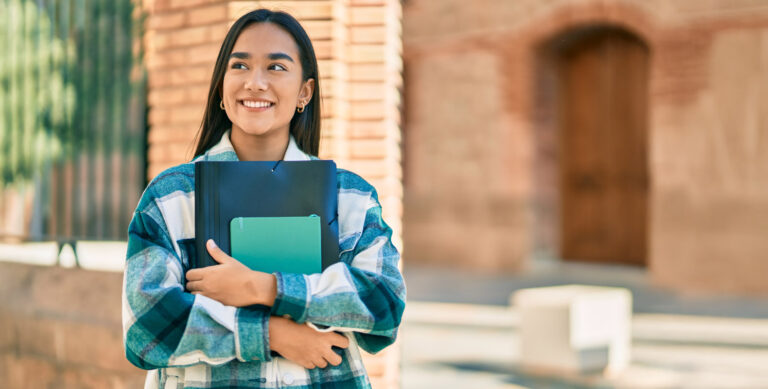 A young woman stands outside, smiling and holding a notebook and a folder. She wears a plaid shirt and has long dark hair. The background features a brick building and an unfocused outdoor setting.