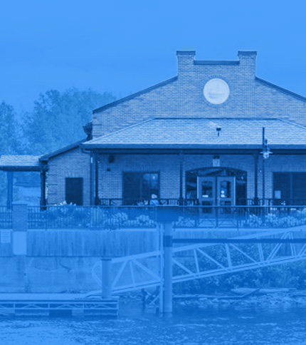 A brick building with a chimney and a large circular sign stands by the water, its entrance bordered by a railing. Trees frame the background. The image, tinted blue, evokes the calm setting of an Education Summit venue.