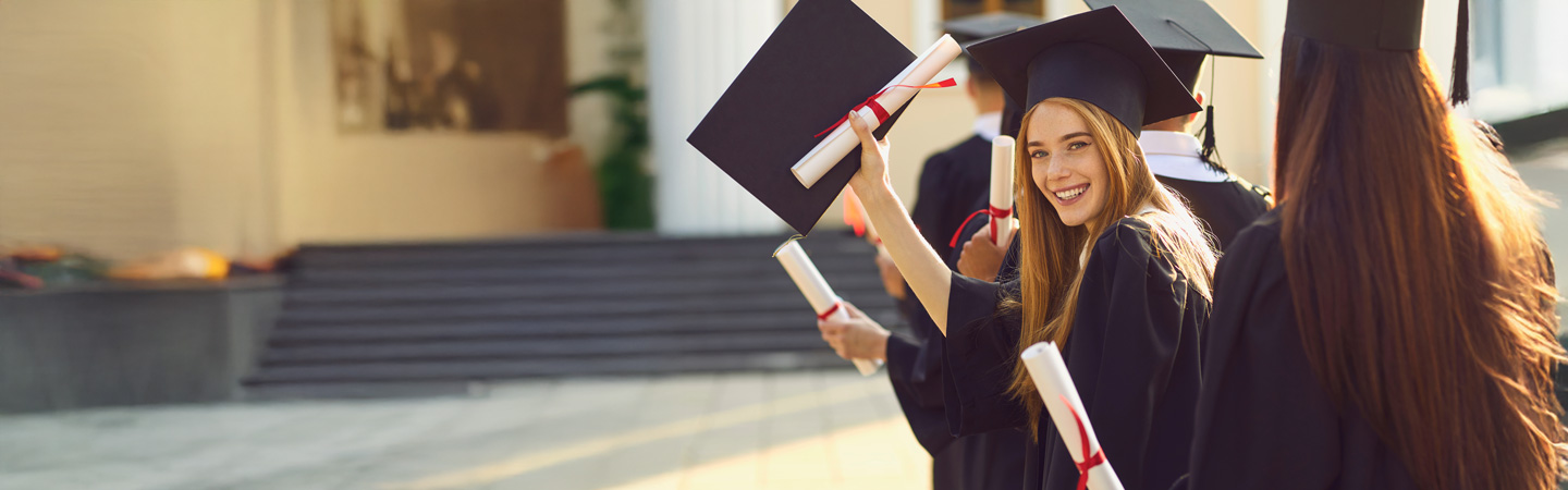 A group of graduates in black caps and gowns stand outside, holding diplomas. One graduate, smiling, is looking back towards the camera. Steps and a building entrance are visible in the background.