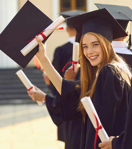 A young woman in a graduation gown and cap smiles while holding up her diploma. Others in similar attire stand in the background, also holding diplomas. They appear to be outside a building, possibly a school or university.