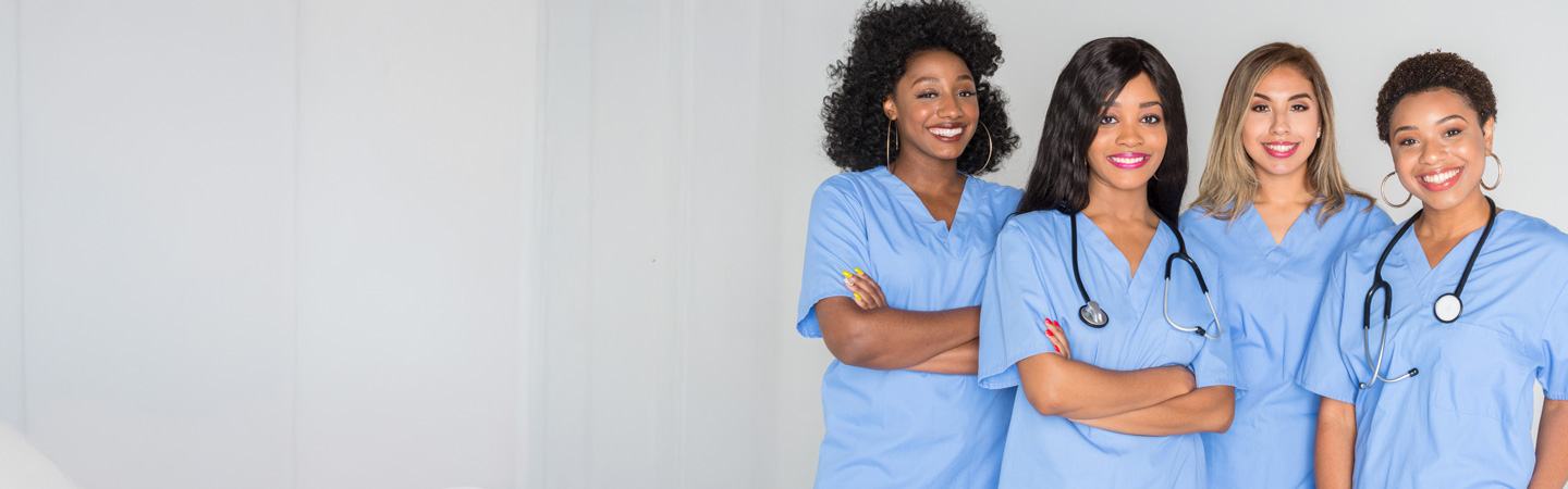 Four healthcare professionals in blue scrubs, smiling and standing together in a clean, clinical setting. Three wear stethoscopes around their necks. The background is bright with soft lighting.
