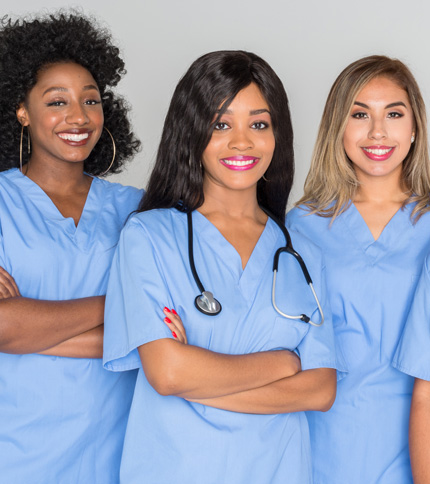 Three women in blue medical scrubs smiling. The woman in the middle has a stethoscope around her neck. They stand side by side, conveying a sense of teamwork and professionalism.