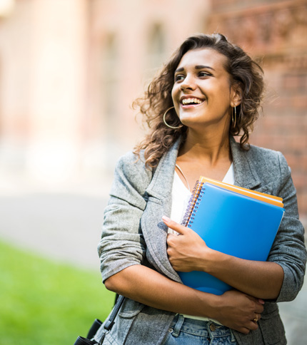 A smiling woman stands outside holding notebooks. She is wearing a gray blazer and hoop earrings, with curly hair. A blurred building and lawn are in the background.