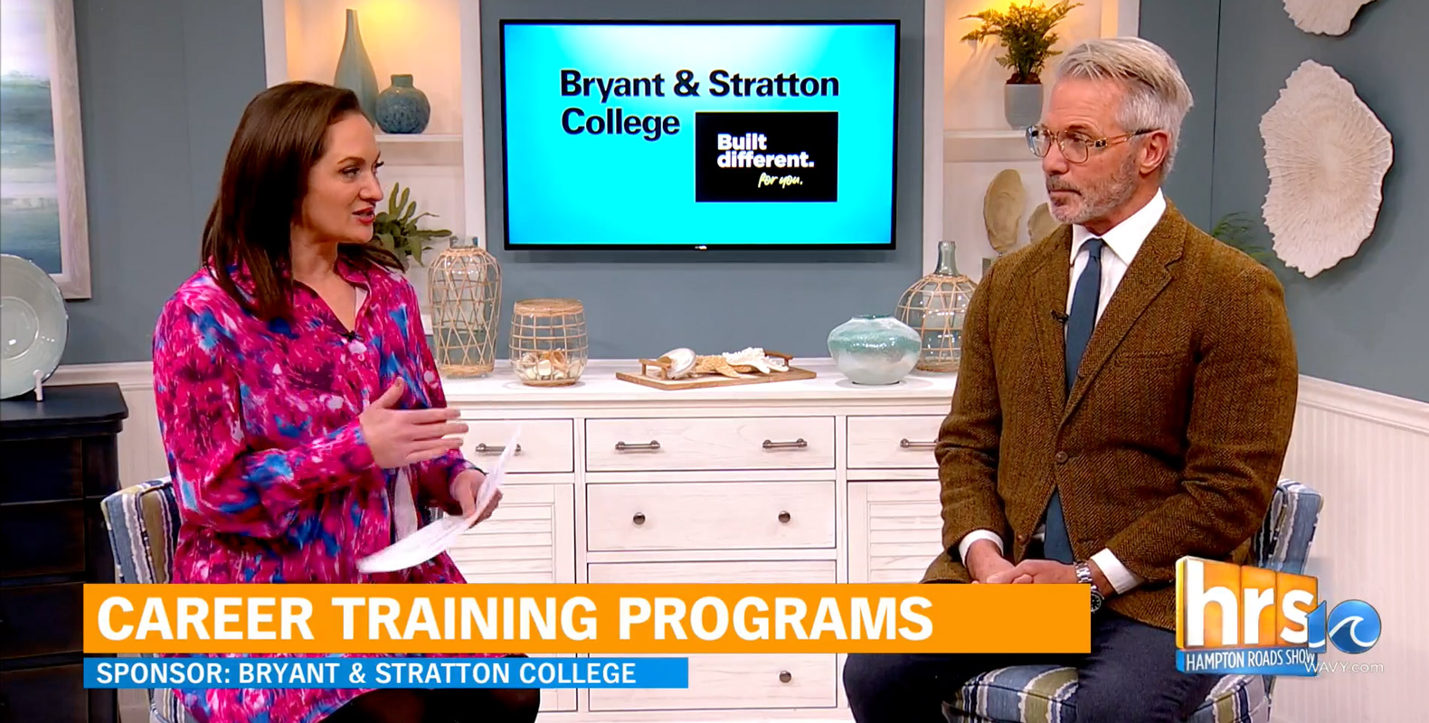 A woman in a pink floral dress interviews a man in a brown blazer on a talk show set. Behind them is a TV displaying "Bryant & Stratton College" and a banner reading "Career Training Programs" appears at the bottom.