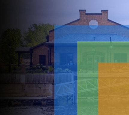 A brick building with a circular window sits by a river. The foreground features overlapping translucent bars in blue, yellow, and orange, partially obscuring the view. Trees and a wooden fence are visible near the structure.
