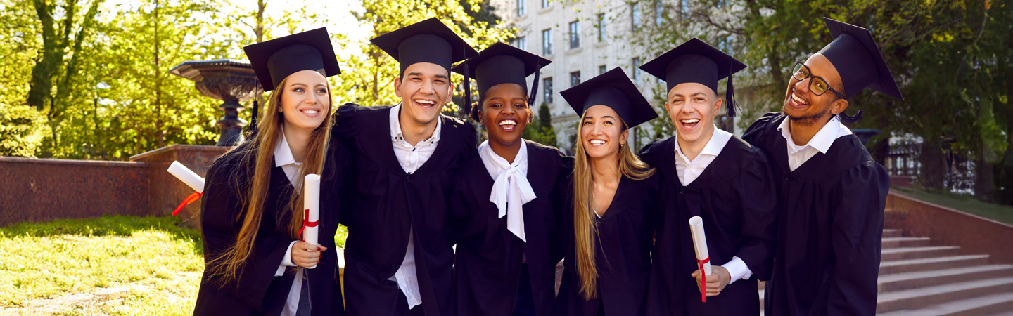 Six graduates in caps and gowns celebrate outdoors, smiling and holding diplomas, with trees and a building in the background.