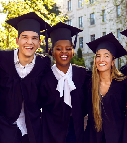 Three graduates wearing black caps and gowns smile broadly. They stand closely together, celebrating outdoors with a building and tree in the background, suggesting a college or university setting.