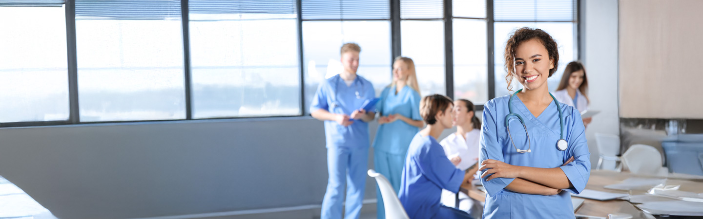 Smiling healthcare worker in scrubs and stethoscope stands with arms crossed in an office. In the background, six medical professionals in similar attire are collaborating near large windows.