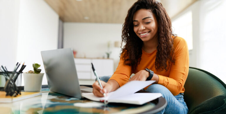 A woman with long curly hair smiles while writing in a notebook at a round glass table. She is wearing an orange sweater and sitting beside a laptop. The room is bright and modern, with some stationery and a small plant on the table.